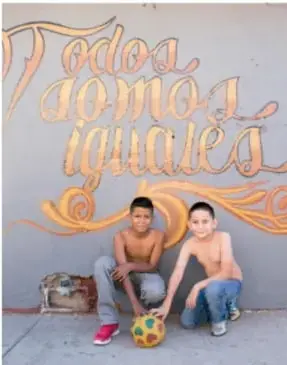 Muro gris con la frase dorada 'Todos somos iguales' y dos niños sonrientes sentados con un balón de fútbol.