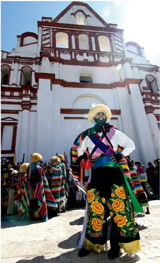 Fotografía de un participante de la danza de los chinelos con traje de colores vivos, sombrero de paja y máscara, de pie frente a una iglesia blanca con ornamentos rojos, mientras otros danzantes y espectadores observan al fondo.