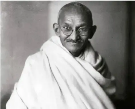Fotografía en blanco y negro de Mahatma Gandhi, con lentes redondos y cubierto con un sari blanco, de perfil ligeramente sonriente.