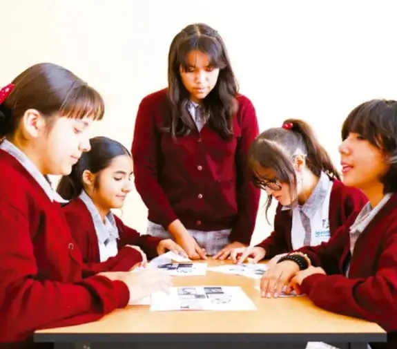 Fotografía de cinco estudiantes de telesecundaria con uniforme rojo trabajando juntos alrededor de una mesa, revisando materiales impresos de su proyecto.
