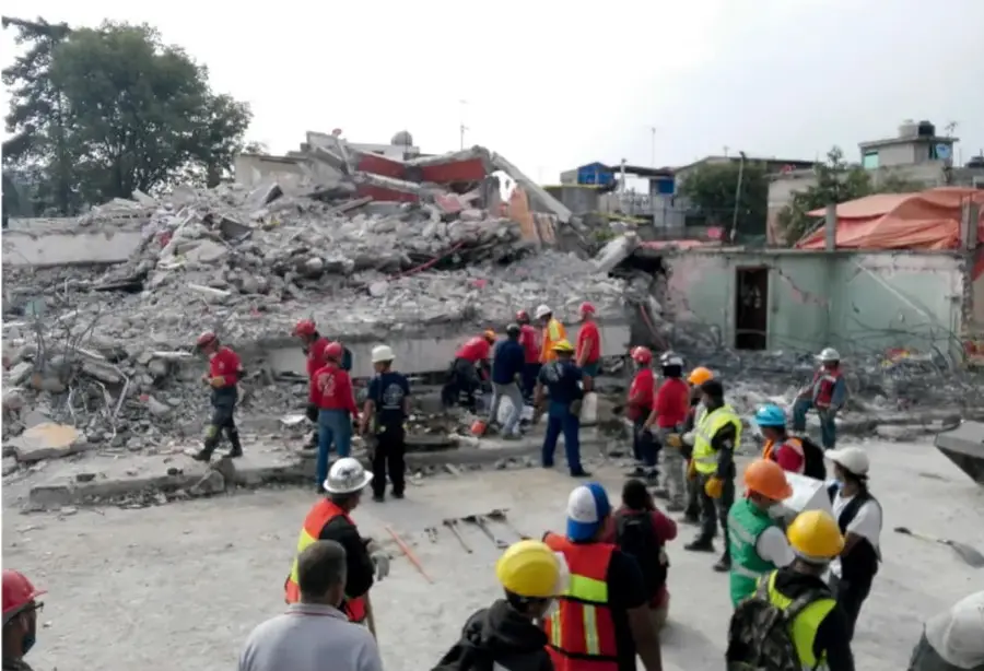 Fotografía de una zona urbana con edificios colapsados y escombros tras un sismo. Se observan rescatistas con cascos y chalecos reflectantes trabajando entre los restos de las construcciones.