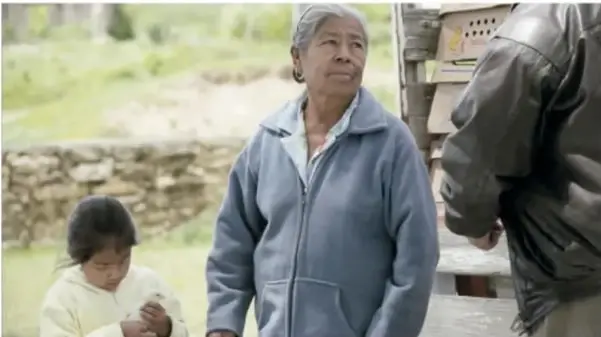 Fotograma del cortometraje: una mujer mayor con suéter azul y una niña indígena observan a un hombre de espaldas.