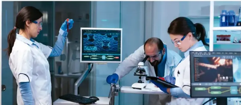 Laboratorio con tres científicos: una mujer con guantes y gafas manipula una muestra, un hombre observa un microscopio y otra mujer revisa una tableta; hay pantallas con datos.