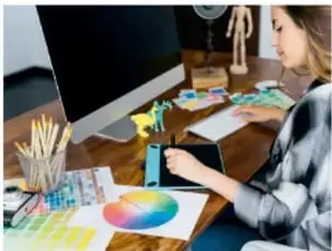 Mujer trabajando en un escritorio con computadora, tablet y muestras de color, representando el ámbito del diseño.