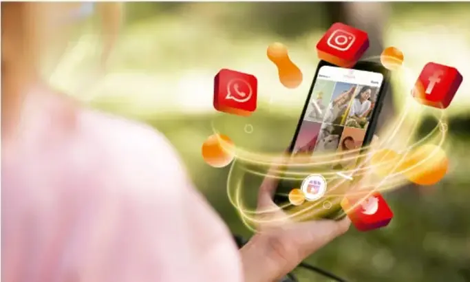 Fotografía de una persona sosteniendo un teléfono inteligente con iconos flotantes de redes sociales (WhatsApp, Instagram, YouTube, etc.) y esferas naranjas, simbolizando la conexión digital.