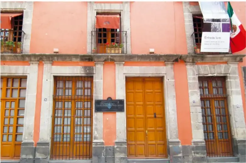 Fachada naranja de la casa colonial con puertas y balcones de madera, escudos de piedra, cartel de entrada y banderas de México; debajo, la leyenda con la dirección.