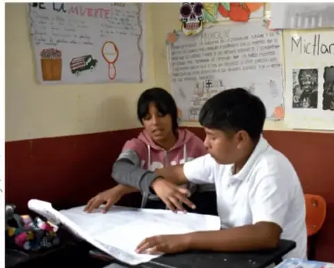 Fotografía de dos estudiantes trabajando juntos sobre una mesa con un pliego de papel, elaborando su periódico mural; al fondo un mural escolar con dibujos y texto.