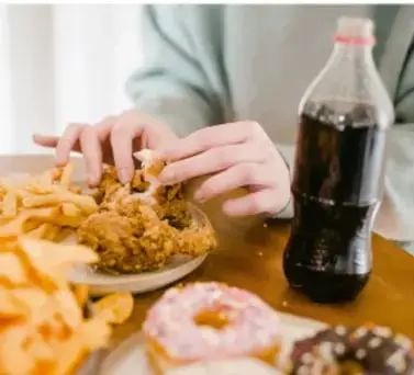 Fotografía de manos tomando frituras y una bebida azucarada sobre una mesa, con alimentos empaquetados y refresco.