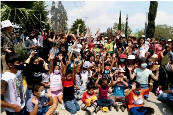 Fotografía de una comunidad reunida al aire libre, con niños, niñas y adultos levantando la mano, en un ambiente de participación colectiva.