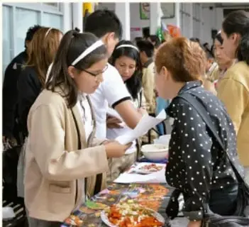 Fotografía de visitantes (adultas y jóvenes) observando las piezas de la exposición mientras una estudiante con diadema les explica.