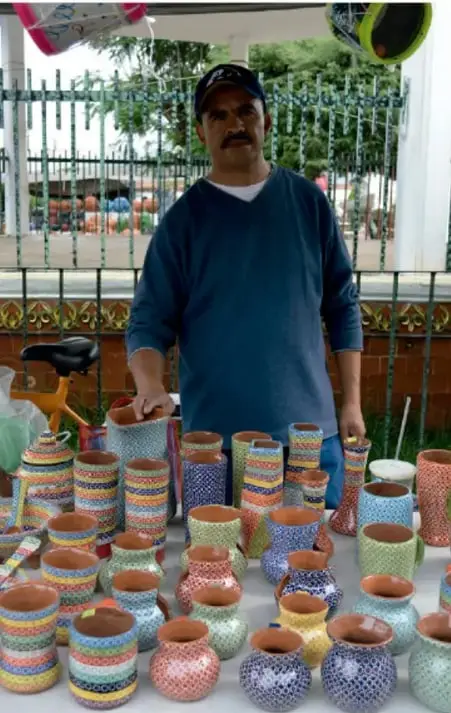 Artesano junto a una gran cantidad de piezas de alfarería coloridas, probablemente en Capula, Michoacán.