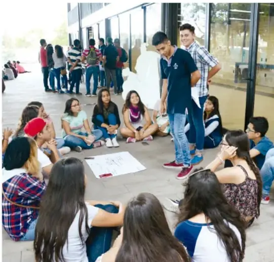 Fotografía de estudiantes de secundaria sentados en círculo en el patio de la escuela conversando en asamblea, con un papelógrafo y un estudiante de pie coordinando.