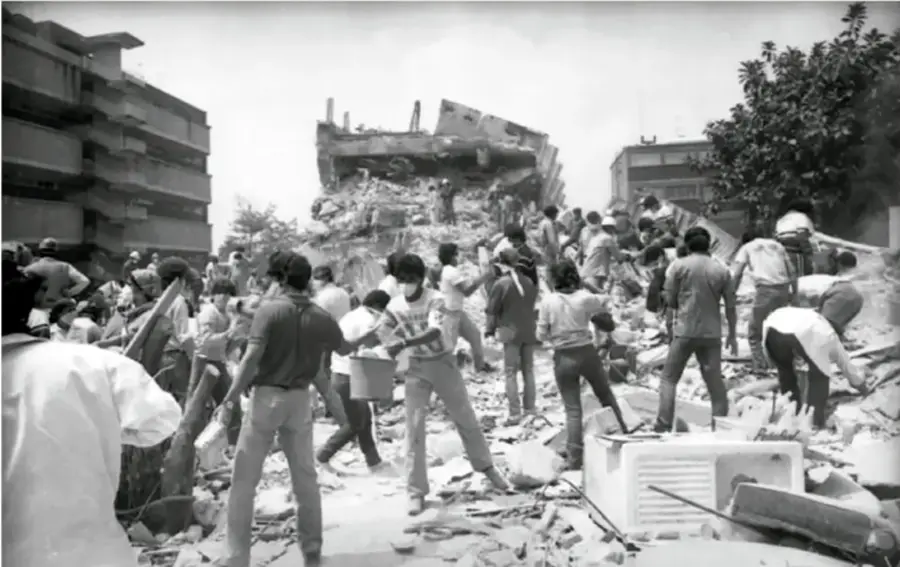Fotografía en blanco y negro de una escena de rescate tras el sismo de 1985 en México. Se observan personas entre escombros de edificios colapsados, algunas con cascos, y una multitud trabajando en labores de rescate. Al fondo, estructuras dañadas y árboles.