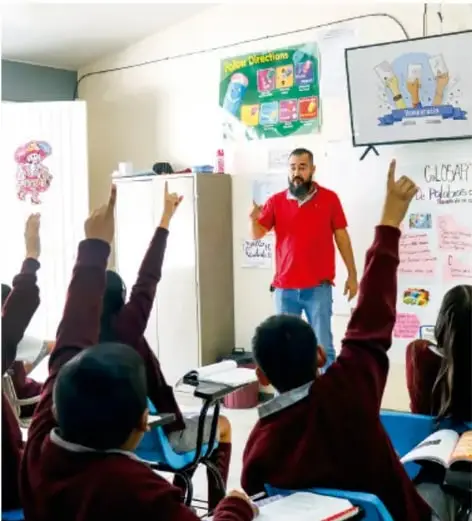 Fotografía de un aula de Telesecundaria: el docente, de camiseta roja, explica frente a un pizarrón con carteles mientras los estudiantes levantan la mano para pedir el turno de palabra, ilustrando la participación democrática en el aula.