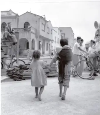 Fotografía en blanco y negro de una calle empedrada con personas caminando, entre ellas una niña y un adulto con carga, en un entorno rural o urbano antiguo.