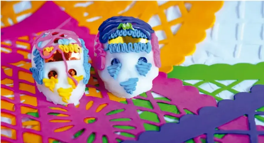 Dos calaveritas decoradas con papel picado de colores (rosa, azul, amarillo, verde y morado) sobre fondo blanco; una tiene detalles azules y la otra detalles amarillos y rojos. Se relacionan con el Día de Muertos.