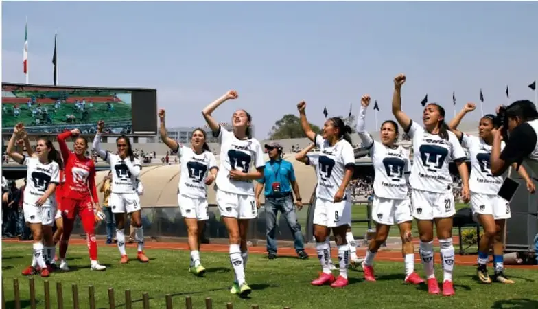 Fotografía de un equipo de fútbol femenil con camisetas blancas celebrando con los brazos en alto en la cancha; se ve el logotipo de la liga en el estadio.