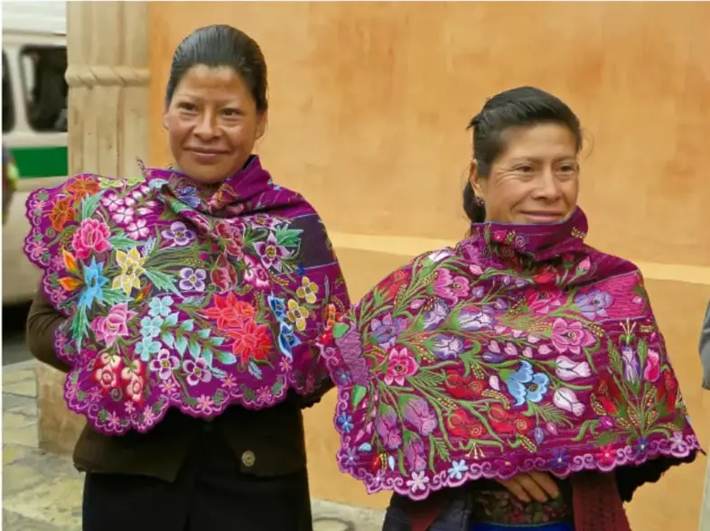 Fotografía de dos mujeres indígenas sonriendo, visten rebozos o chales bordados con motivos florales de colores vivos (rosa, morado, verde), en un espacio urbano con muro ocre al fondo.