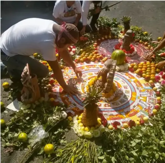 Fotografía de personas inclinadas preparando una ofrenda o alfombra ritual circular en el suelo, elaborada con frutas, plantas y objetos ceremoniales; correspondiente a la imagen referenciada como page=0,bbox=[151,478,681,1003].