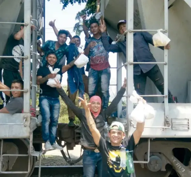 Grupo de jóvenes migrantes sonriendo y saludando desde las escaleras y el techo de un tren de carga, algunos con bolsas blancas de comida en las manos.