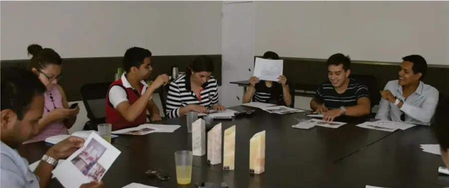 Fotografía de un grupo de jóvenes y adultas sentados alrededor de una mesa larga en una reunión de trabajo, revisando documentos y libretas; algunas personas sostienen hojas y conversan. Crédito: Gimenna Schiaffini Rosales, Puebla.