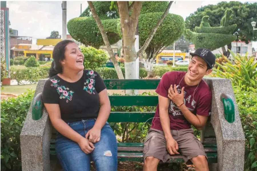 Una joven y un joven conversan y ríen sentados en un banco de un parque con árboles esculpidos (topiaria) de fondo.