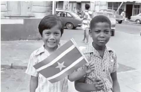 Fotografía en blanco y negro de dos niños sosteniendo la bandera de Surinam en una calle.