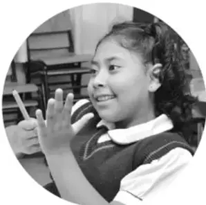 Fotografía en blanco y negro de una niña sonriente en un salón de clases, con las manos levantadas.