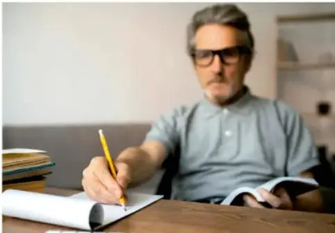 Fotografía de un hombre con lentes escribiendo con lápiz en un cuaderno, sentado en una mesa de madera con libros.