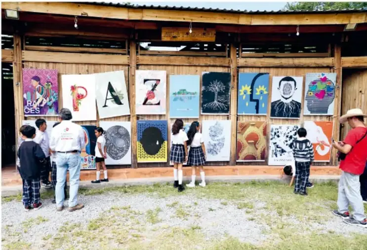 Fotografía de estudiantes de secundaria observando una muestra de murales y carteleras colgados en la fachada de un edificio escolar de madera; varios carteles forman la palabra PAZ y hay imágenes de personas, árboles y símbolos de convivencia.
