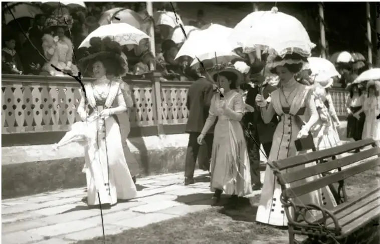 Fotografía en blanco y negro de mujeres de la élite porfiriana paseando con vestidos largos, sombreros y sombrillas, junto a una banca y una balaustrada.