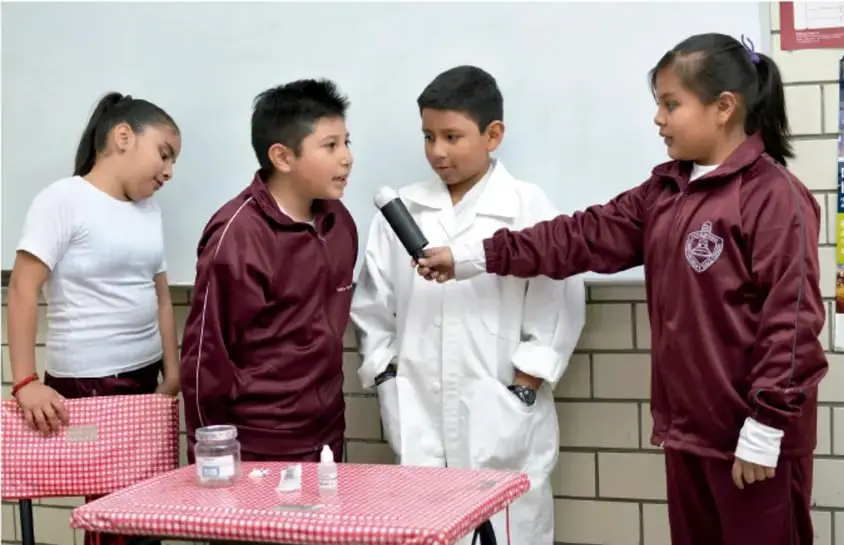 Fotografía de cuatro estudiantes de primaria alrededor de una mesa con mantel a cuadros rojos; uno de ellos sostiene un micrófono y parece entrevistar a otro, en un patio escolar.