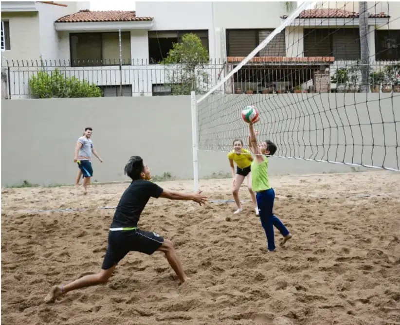 Fotografía de jóvenes jugando voleibol de playa en una cancha de arena, con red y viviendas al fondo; ilustra la actividad física al aire libre.