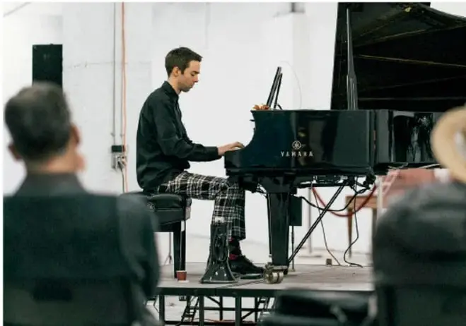 Fotografía de un pianista tocando un piano de cola Yamaha frente a un grupo de personas, en un espacio amplio tipo taller.
