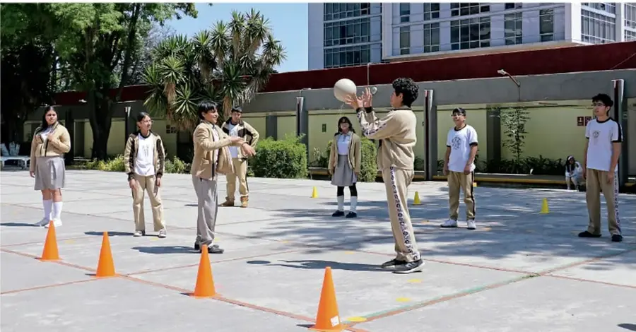 Fotografía de estudiantes de telesecundaria jugando con una pelota en una cancha escolar, con conos naranjas que delimitan las bases y un estudiante a punto de lanzar la pelota.