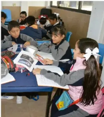 Fotografía de estudiantes de primaria trabajando en equipo en un aula, con libros y cuadernos sobre la mesa.