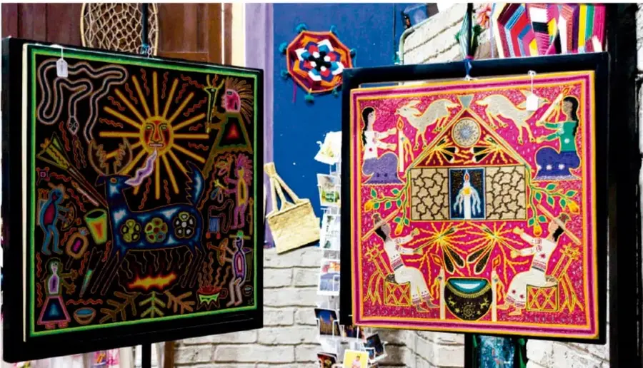 Fotografía de un mercado o calle con obras de arte popular expuestas: dos piezas textiles/marco con motivos coloridos (una de fondo negro con sol y figuras, otra de fondo rosa con figuras humanas, animales y casita), además de accesorios tejidos en el muro azul.