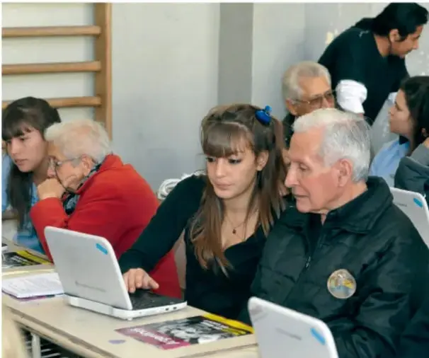 Fotografía de un taller comunitario donde personas mayores y jóvenes trabajan juntas frente a laptops, ilustrando la interacción intergeneracional y el uso de herramientas tecnológicas en la comunidad.
