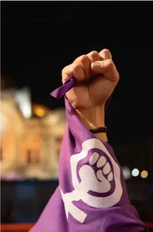 Fotografía de un puño levantado sosteniendo una tela morada con un símbolo de puño blanco, en un contexto de manifestación o protesta social.