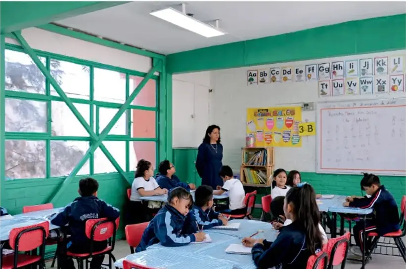 Fotografía de un aula con paredes verdes: una maestra atiende a sus alumnas y alumnos sentados en mesas redondas con manteles azules; al fondo hay carteleras con el abecedario (Aa, Bb, Cc...), el rótulo 3:B y una pizarra blanca con el tema del día.