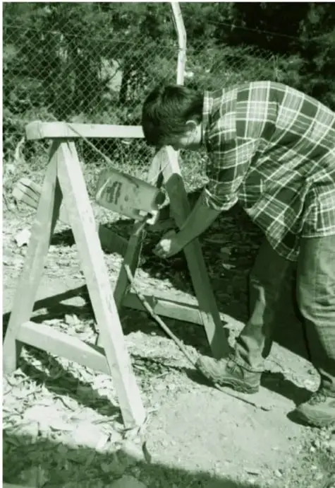 Fotografía en tono verde de una persona adulta instalando el recipiente (botella) en la estructura de madera del tippy tap, junto a una reja, al aire libre.