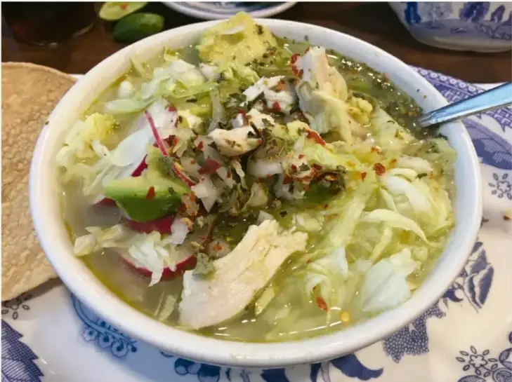 Fotografía de un cuenco de caldo verde con pollo, lechuga, rábanos, aguacate y chile, acompañado de tortillas tostadas sobre un mantel azul y blanco; ilustra una comida típica representativa.