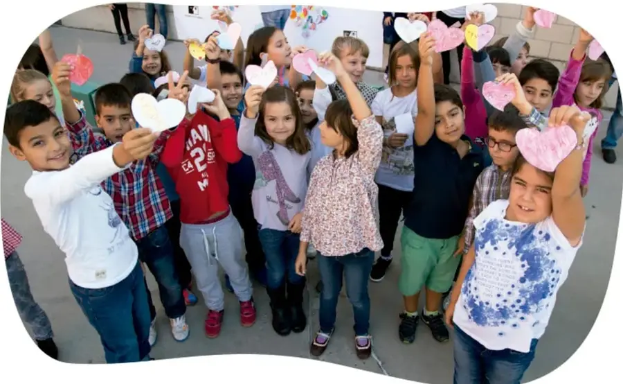 Fotografía de un grupo grande de niños y niñas de primaria al aire libre, sonriendo y levantando corazones de cartulina de colores (blancos, rosas, amarillos y rojos).