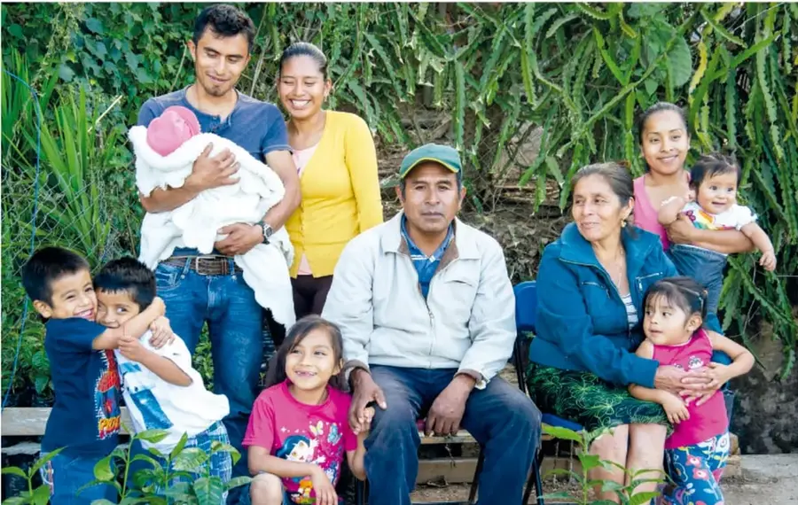 Fotografía de una familia numerosa al aire libre frente a plantas verdes: un papá carga a un bebé con gorrito rosa, hay dos niños abrazados, una niña sonriente, un señor sentado con gorra, una señora mayor (abuela) y una joven que carga a una bebé, con otra niña pequeña sentada.