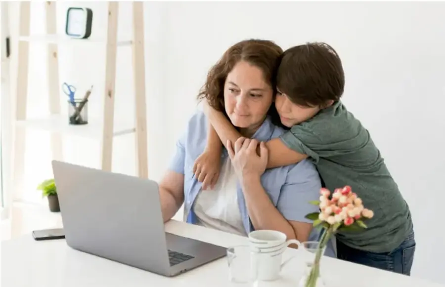 Fotografía de una mujer sentada frente a una laptop abrazando a un niño que se acerca por detrás; sobre la mesa hay tazas y flores. Representa el apoyo emocional en familia.
