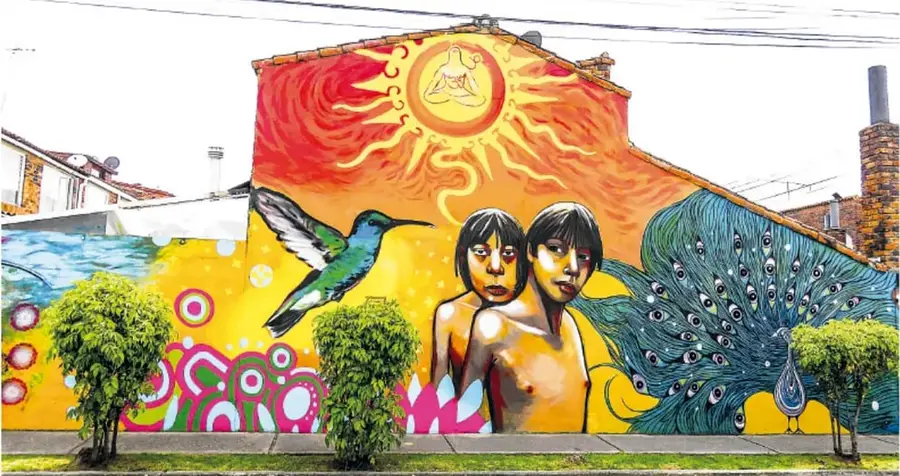 Mural urbano a todo color sobre una fachada: un sol radiante, niños indígenas en el centro, un colibrí volando y un pavo real con el plumaje extendido, entre formas abstractas de colores cálidos.
