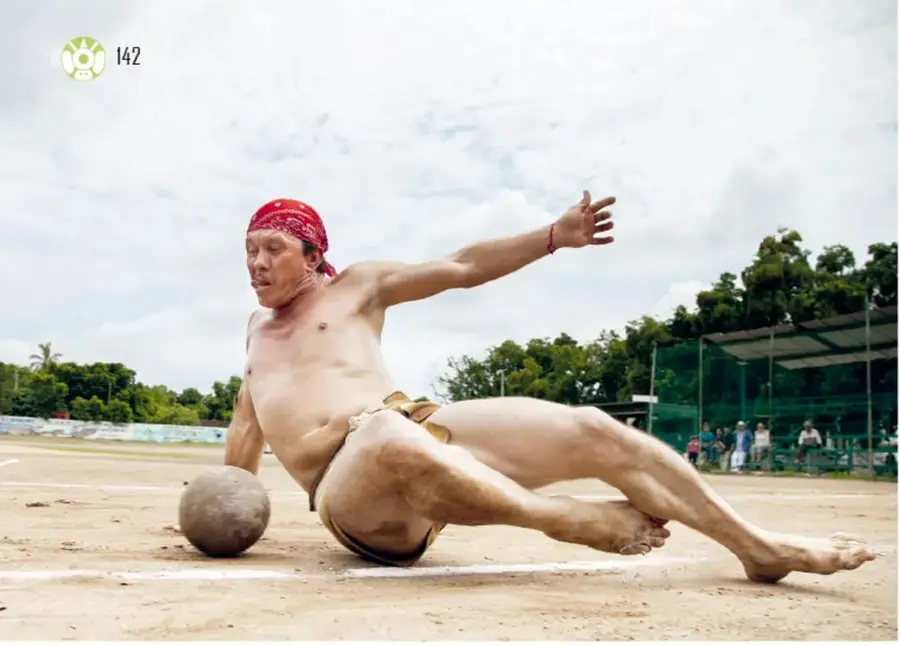Jugador de ulama descalzo, con taparrabo y paño rojo en la cabeza, golpeando con la cadera una pelota de hule en una cancha de tierra; al fondo hay público y árboles.