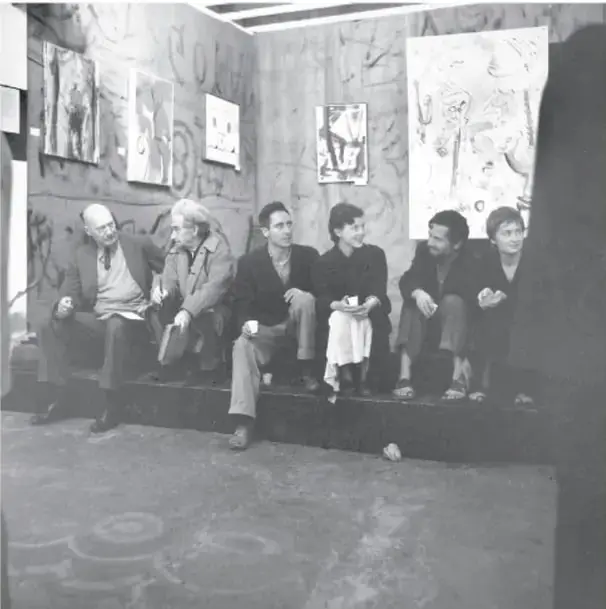 Fotografía en blanco y negro de un grupo de personas sentadas en lo que parece ser una galería o espacio cultural con dibujos en las paredes.