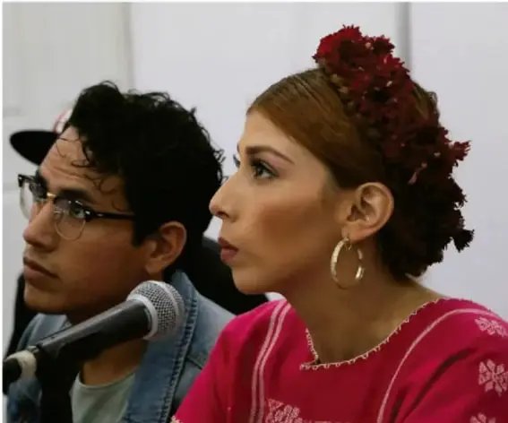 Fotografía de dos jóvenes; uno con lentes y gorra sostiene un micrófono y la otra lleva prendas y adornos tradicionales (flores en el cabello, aretes grandes, blusa bordada), ilustrando la mezcla de identidades contemporáneas y tradicionales.