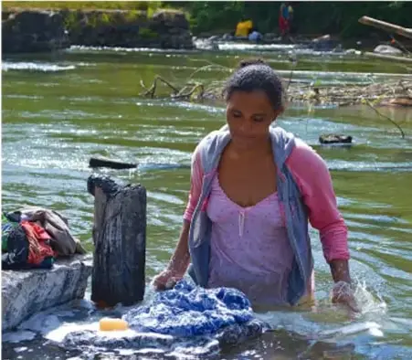 Fotografía de una mujer lavando ropa en un río, ilustrando el trabajo cotidiano y las relaciones de desigualdad en la vida diaria.